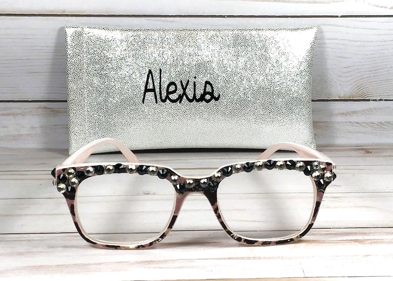 May include: Eyeglasses with a patterned frame and embellished with dark rhinestones. The glasses are paired with a silver pouch with the name "Alexia" in black script.
