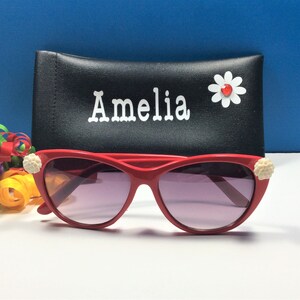 May include: Red cat-eye sunglasses with white flower embellishments on the sides. The sunglasses are in a black case with the name "Amelia" written on it.