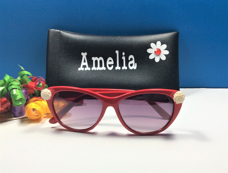 May include: Red cat-eye sunglasses with white flower embellishments on the sides. The sunglasses are in a black case with the name "Amelia" written on it.