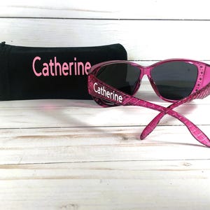 May include: Pink sunglasses with dark lenses and a matching black case. The sunglasses have a textured pattern on the frames and the name "Catherine" is printed on the frames and case.