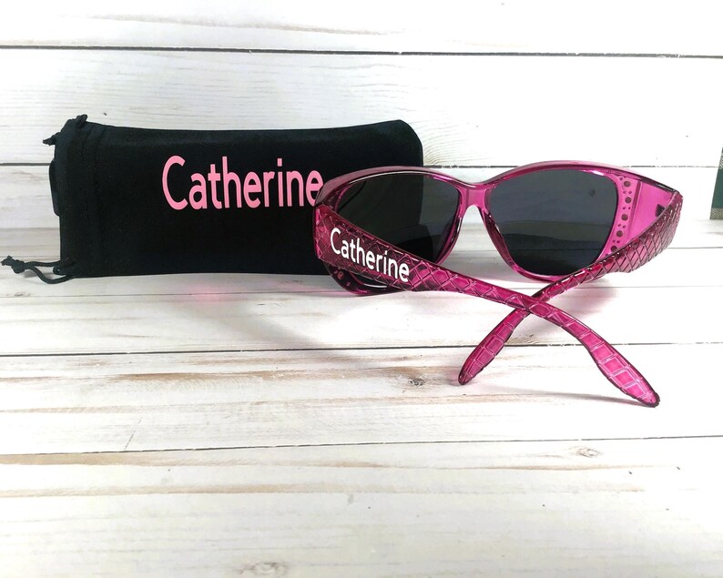 May include: Pink sunglasses with dark lenses and a matching black case. The sunglasses have a textured pattern on the frames and the name "Catherine" is printed on the frames and case.