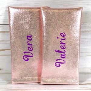 Pu&ograve; includere: Due pochette color oro rosa con una superficie strutturata e puntinata. La parola "Vera" &egrave; scritta in corsivo viola sulla pochette di sinistra e "Valerie" &egrave; scritta sulla pochette di destra. Le pochette sono rettangolari e sembrano essere realizzate in un materiale lucido.