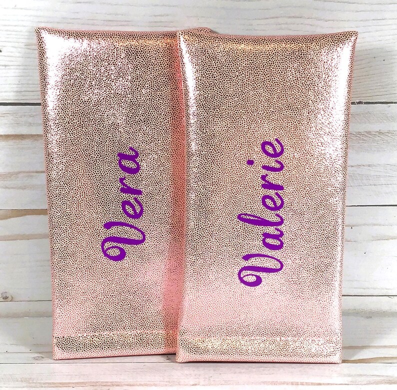 Pu&ograve; includere: Due pochette color oro rosa con una superficie strutturata e puntinata. La parola "Vera" &egrave; scritta in corsivo viola sulla pochette di sinistra e "Valerie" &egrave; scritta sulla pochette di destra. Le pochette sono rettangolari e sembrano essere realizzate in un materiale lucido.