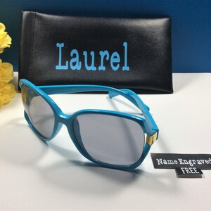May include: Blue sunglasses with a black case. The case has the name "Laurel" printed on it. A tag is attached to the sunglasses that says "Name Engraved FREE".