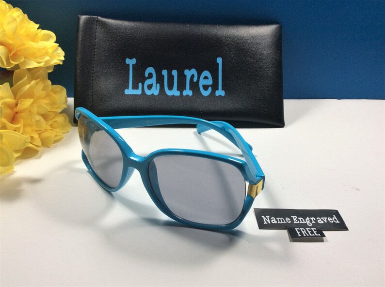 May include: Blue sunglasses with a black case. The case has the name "Laurel" printed on it. A tag is attached to the sunglasses that says "Name Engraved FREE".