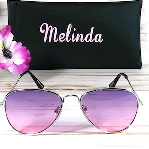 Peut inclure: Lunettes de soleil aviateur argent&eacute;es avec des verres d&eacute;grad&eacute;s rose &agrave; violet. Les lunettes sont sur une surface blanche avec un &eacute;tui noir derri&egrave;re elles. L'&eacute;tui porte le nom "Melinda" brod&eacute; en rose.