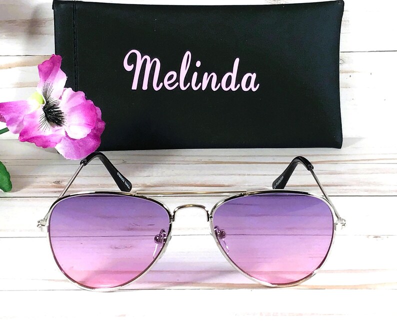 Peut inclure: Lunettes de soleil aviateur argent&eacute;es avec des verres d&eacute;grad&eacute;s rose &agrave; violet. Les lunettes sont sur une surface blanche avec un &eacute;tui noir derri&egrave;re elles. L'&eacute;tui porte le nom "Melinda" brod&eacute; en rose.