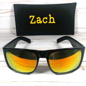 Puede incluir: Gafas de sol negras con lentes espejadas naranjas y una funda negra. La funda tiene el nombre "Zach" en letras amarillas. Las gafas tienen una montura rectangular cl&aacute;sica.