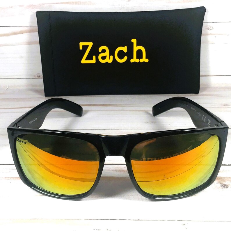 Puede incluir: Gafas de sol negras con lentes espejadas naranjas y una funda negra. La funda tiene el nombre "Zach" en letras amarillas. Las gafas tienen una montura rectangular cl&aacute;sica.
