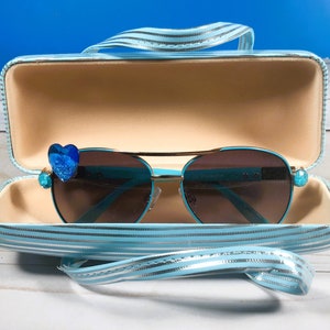 May include: Turquoise aviator sunglasses with brown lenses, featuring blue heart and jewel embellishments. The sunglasses are presented in a matching striped case with a handle.