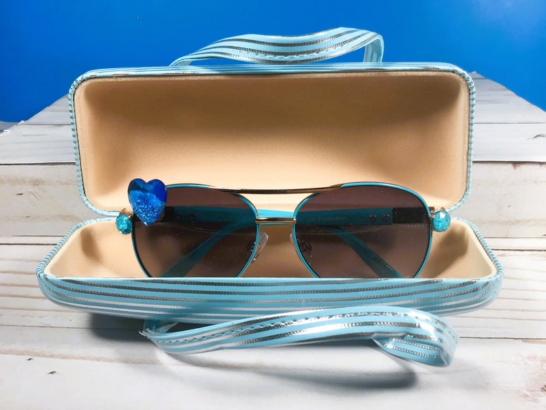 May include: Turquoise aviator sunglasses with brown lenses, featuring blue heart and jewel embellishments. The sunglasses are presented in a matching striped case with a handle.