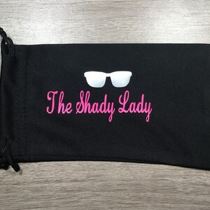 May include: Black drawstring bag with a white pair of sunglasses and the text "The Shady Lady" in pink.