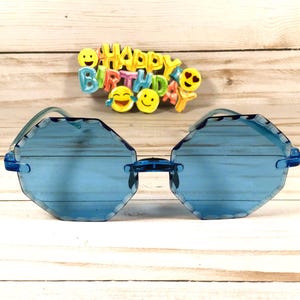 May include: Blue octagon shaped sunglasses with a light blue lens. The sunglasses are on a light wood background with a colorful "Happy Birthday" sign above them.