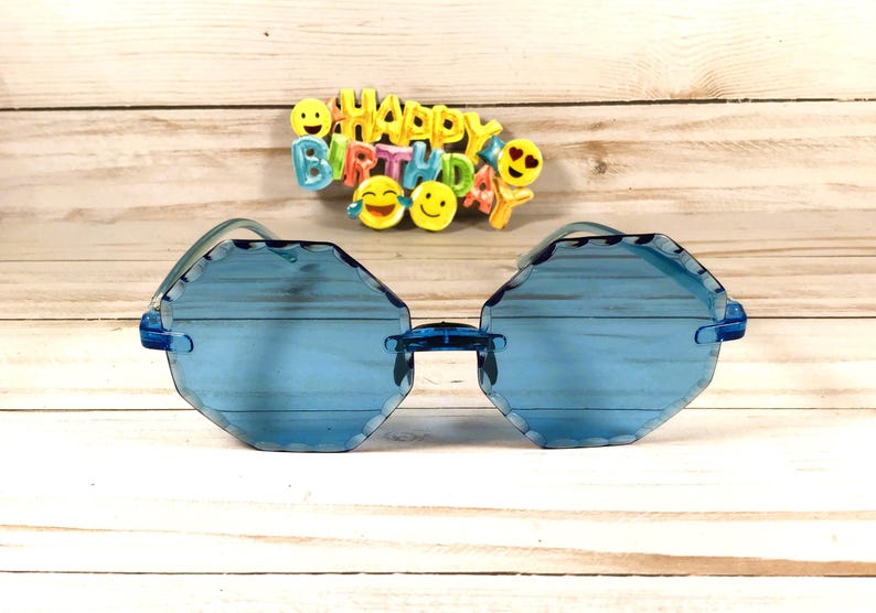 May include: Blue octagon shaped sunglasses with a light blue lens. The sunglasses are on a light wood background with a colorful "Happy Birthday" sign above them.