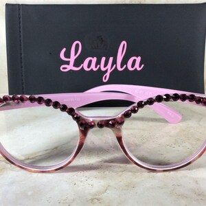 K&ouml;nnte beinhalten: Rosa Brille mit dunkelroten Strasssteinen am oberen Rand der Fassung. Die Gl&auml;ser sind klar. Hinter der Brille befindet sich ein schwarzes Etui mit dem Namen "Layla" in rosa Schrift.
