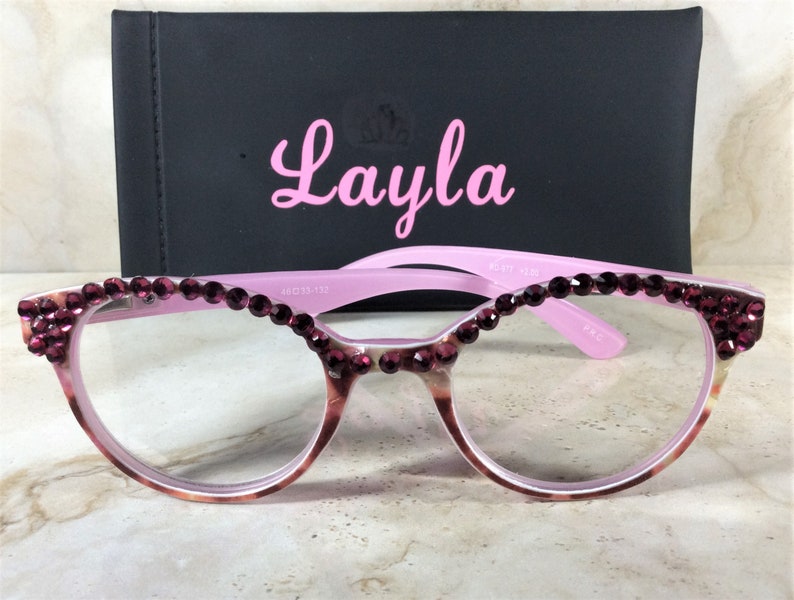 K&ouml;nnte beinhalten: Rosa Brille mit dunkelroten Strasssteinen am oberen Rand der Fassung. Die Gl&auml;ser sind klar. Hinter der Brille befindet sich ein schwarzes Etui mit dem Namen "Layla" in rosa Schrift.