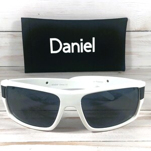 Pu&ograve; includere: Occhiali da sole bianchi con lenti scure e custodia nera con il nome "Daniel" in bianco. Gli occhiali hanno una spessa montatura bianca e una fascia nera sulle aste.