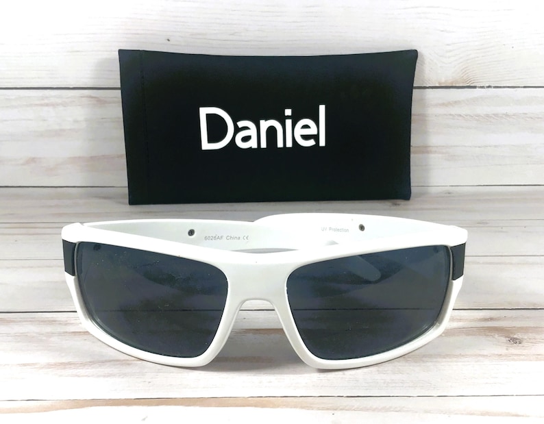 Pu&ograve; includere: Occhiali da sole bianchi con lenti scure e custodia nera con il nome "Daniel" in bianco. Gli occhiali hanno una spessa montatura bianca e una fascia nera sulle aste.