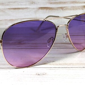 May include: Aviator sunglasses with a gold frame and purple gradient lenses. The lenses fade from a dark purple to a lighter pink. The sunglasses are on a light wooden surface. The frame is a classic design.