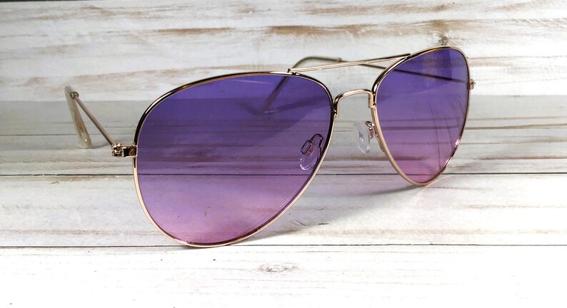 May include: Aviator sunglasses with a gold frame and purple gradient lenses. The lenses fade from a dark purple to a lighter pink. The sunglasses are on a light wooden surface. The frame is a classic design.