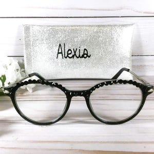 Puede incluir: Gafas negras con borde de montura festoneado y lentes transparentes, colocadas frente a un estuche rectangular de purpurina plateada con el nombre Alexia bordado en hilo negro en la parte frontal del estuche.