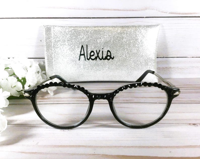 Puede incluir: Gafas negras con borde de montura festoneado y lentes transparentes, colocadas frente a un estuche rectangular de purpurina plateada con el nombre Alexia bordado en hilo negro en la parte frontal del estuche.