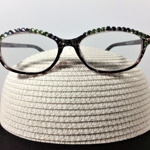 May include: Black-framed eyeglasses with clear lenses, adorned with green and purple rhinestones along the top rim. The glasses rest on a white straw hat with a black band, creating a stylish accessory combination.