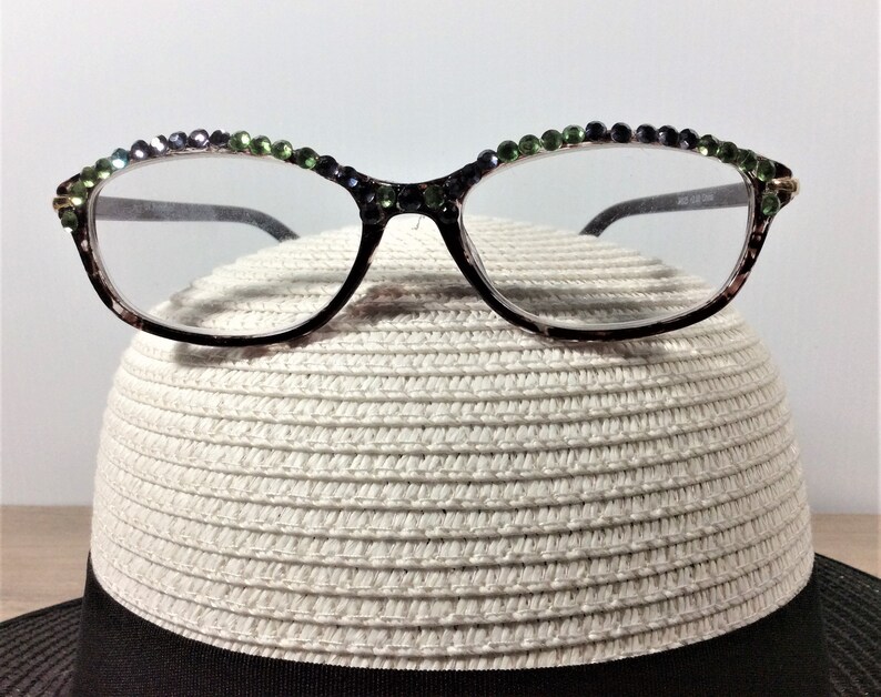 May include: Black-framed eyeglasses with clear lenses, adorned with green and purple rhinestones along the top rim. The glasses rest on a white straw hat with a black band, creating a stylish accessory combination.