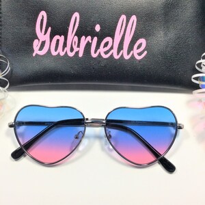 May include: Heart-shaped sunglasses with a blue to pink gradient lens. The sunglasses are on a white surface with a black case that says "Gabrielle" in pink lettering.