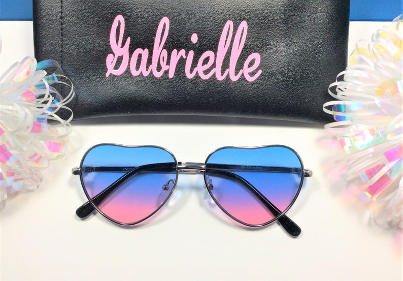 May include: Heart-shaped sunglasses with a blue to pink gradient lens. The sunglasses are on a white surface with a black case that says "Gabrielle" in pink lettering.