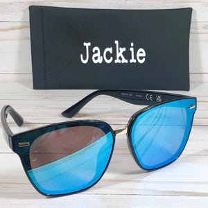 May include: Black sunglasses with blue mirrored lenses and a gold accent on the bridge. The sunglasses have the text "Jackie" on the case.