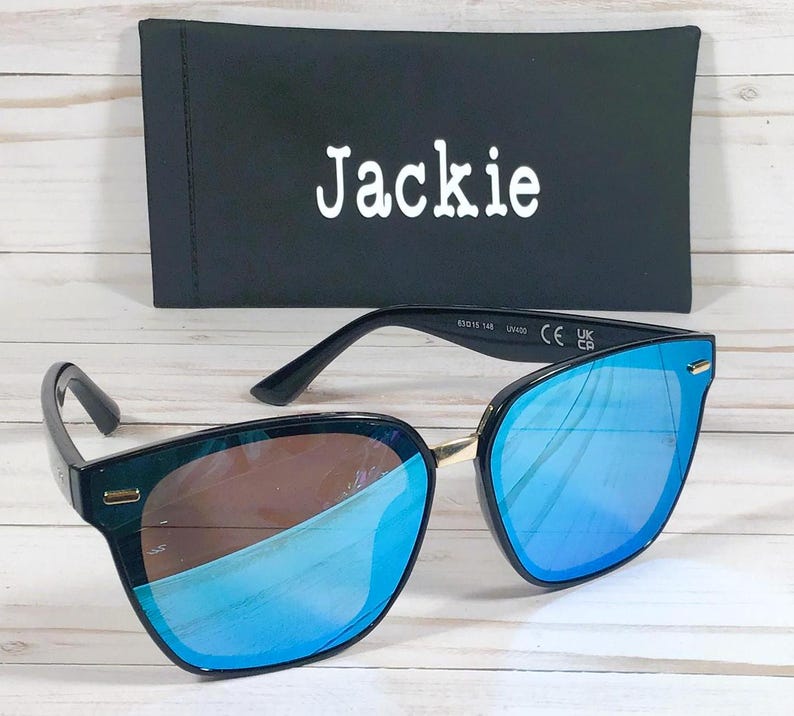 May include: Black sunglasses with blue mirrored lenses and a gold accent on the bridge. The sunglasses have the text "Jackie" on the case.