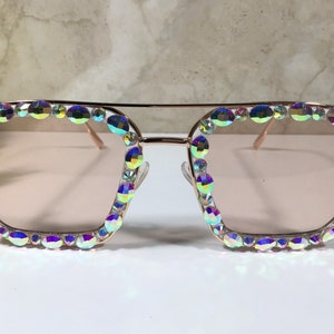 Peut inclure: Lunettes de soleil en or rose avec une monture carr&eacute;e et des strass iris&eacute;s autour des verres.