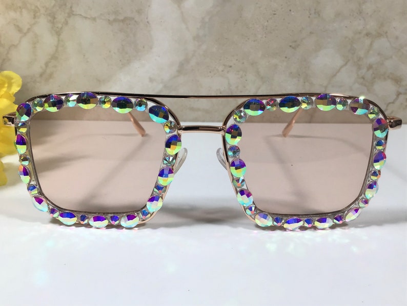 Peut inclure: Lunettes de soleil en or rose avec une monture carr&eacute;e et des strass iris&eacute;s autour des verres.