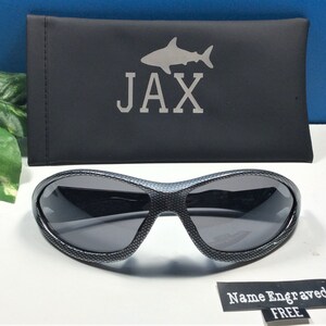 May include: Black sunglasses with a carbon fiber pattern and a gray lens. The sunglasses come with a black carrying case with a shark silhouette and the name "JAX" engraved on it. The text "Name Engraved FREE" is also visible in the image.