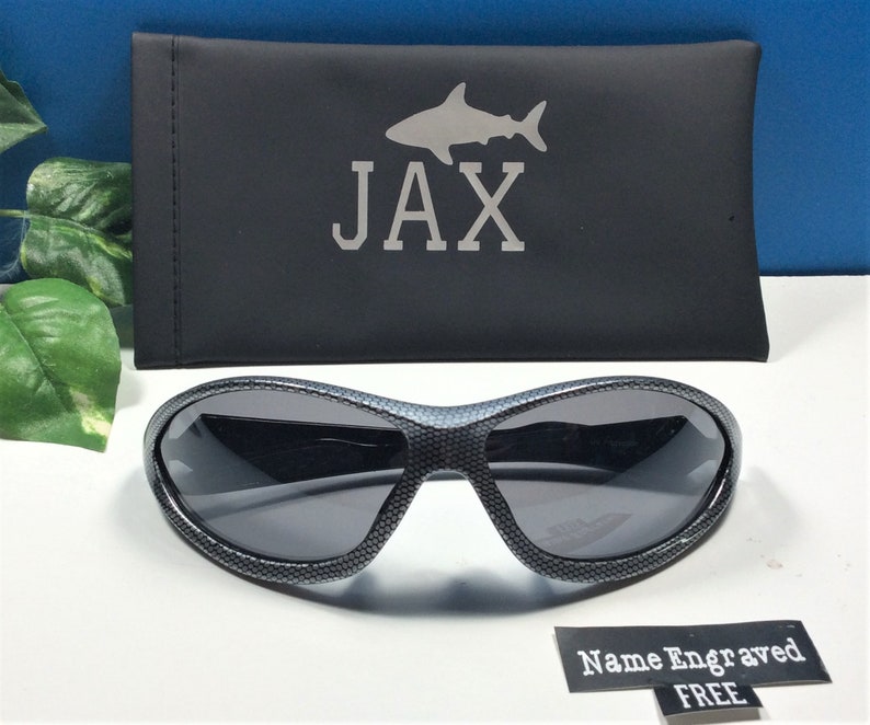 May include: Black sunglasses with a carbon fiber pattern and a gray lens. The sunglasses come with a black carrying case with a shark silhouette and the name "JAX" engraved on it. The text "Name Engraved FREE" is also visible in the image.