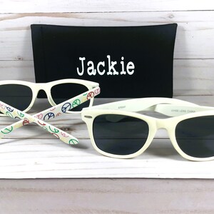 Op de afbeelding: Witte zonnebril met zwarte glazen en een peace-tekenpatroon op de pootjes. De zonnebril wordt geleverd met een zwarte etui met de naam "Jackie" erop geborduurd.