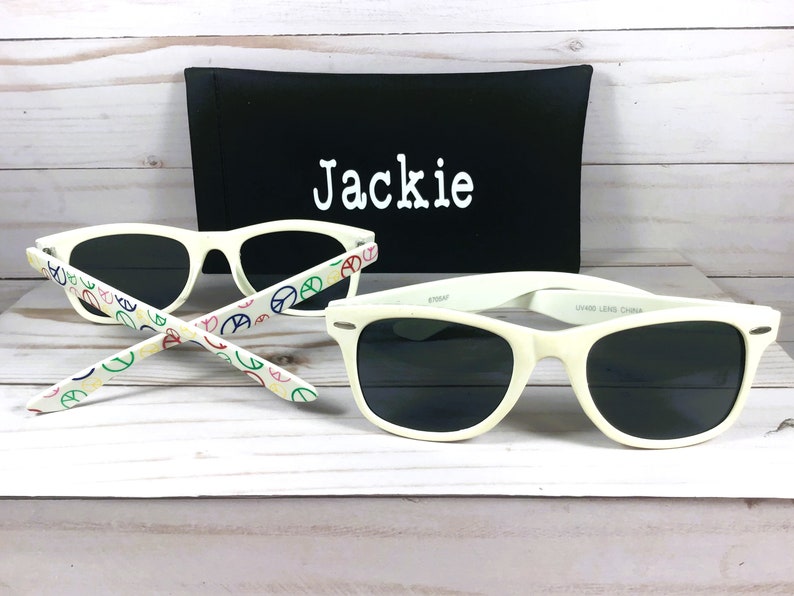 Op de afbeelding: Witte zonnebril met zwarte glazen en een peace-tekenpatroon op de pootjes. De zonnebril wordt geleverd met een zwarte etui met de naam "Jackie" erop geborduurd.