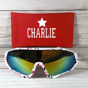 May include: White sunglasses with a pink and black paint splatter pattern and reflective blue-yellow lenses. Behind the sunglasses is a rectangular red case featuring a white star icon above the text CHARLIE in white capital letters.