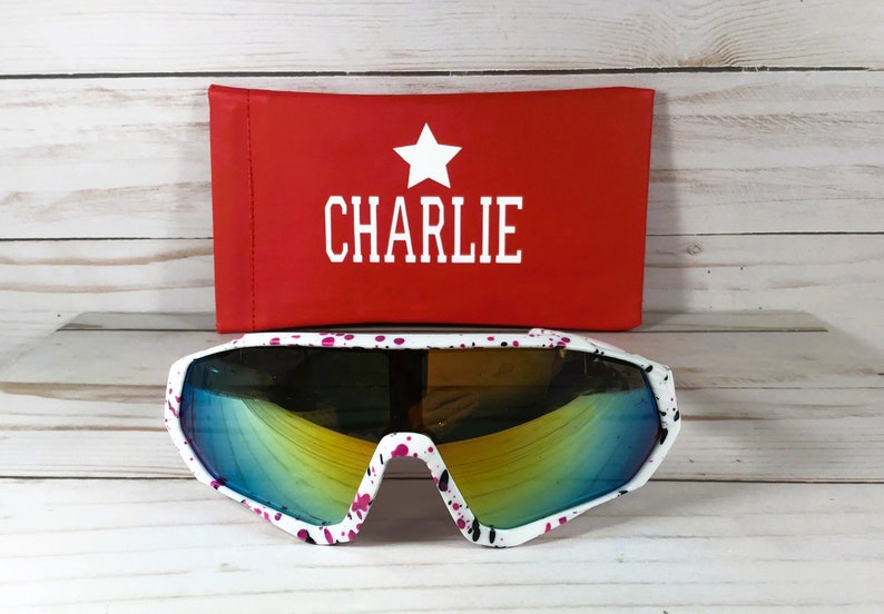 May include: White sunglasses with a pink and black paint splatter pattern and reflective blue-yellow lenses. Behind the sunglasses is a rectangular red case featuring a white star icon above the text CHARLIE in white capital letters.