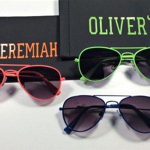 May include: Three pairs of aviator sunglasses in different colors: red, green, and blue. Each pair has a black case with a name on it: Jeremiah, Oliver, and a dinosaur graphic.