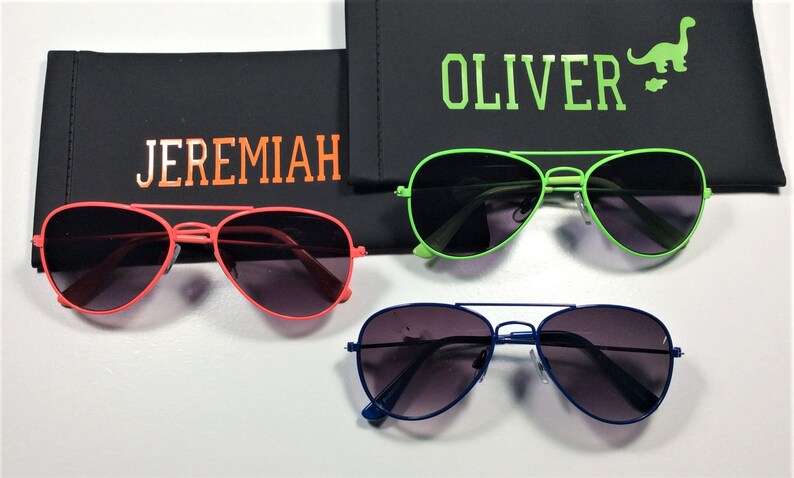 May include: Three pairs of aviator sunglasses in different colors: red, green, and blue. Each pair has a black case with a name on it: Jeremiah, Oliver, and a dinosaur graphic.