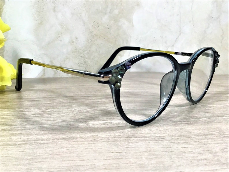 K&ouml;nnte beinhalten: Schwarz umrandete Brille mit klaren Gl&auml;sern und runder Rahmenform. Die &auml;u&szlig;eren Ecken der Rahmen sind mit kleinen, dunklen, kreisf&ouml;rmigen Ziersteinen verziert. Die B&uuml;gel sind aus goldfarbenem Metall mit schwarzen Enden gefertigt.