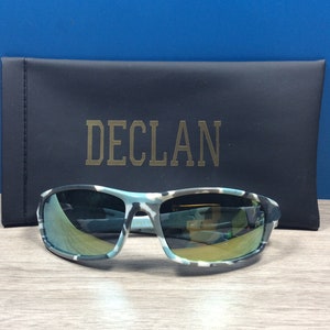 May include: Camouflage patterned sunglasses with a mirrored lens. The sunglasses are in a black case with the word "DECL&Aacute;N" printed in gold.