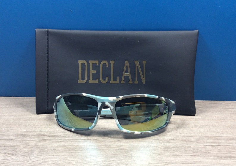 May include: Camouflage patterned sunglasses with a mirrored lens. The sunglasses are in a black case with the word "DECL&Aacute;N" printed in gold.