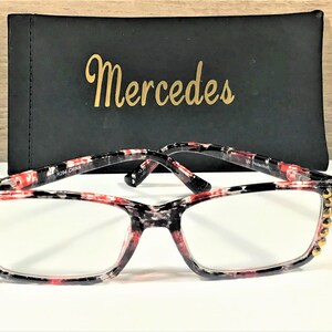 May include: Reading glasses with a black, red, and white patterned frame and clear lenses. The frame has gold-colored embellishments on the corners. A black case with the name "Mercedes" in gold lettering is behind the glasses.