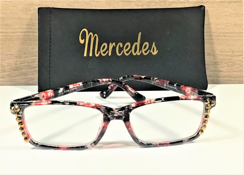May include: Reading glasses with a black, red, and white patterned frame and clear lenses. The frame has gold-colored embellishments on the corners. A black case with the name "Mercedes" in gold lettering is behind the glasses.