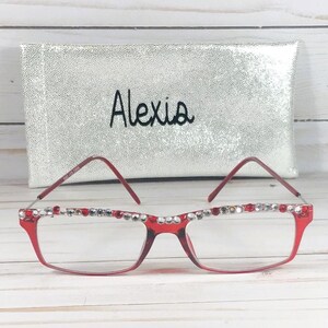 May include: Red-framed eyeglasses with clear lenses, embellished with rhinestones along the top rim. A silver pouch with the name "Alexis" embroidered on it is in the background. The glasses have a rectangular shape.
