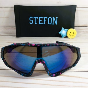 Puede incluir: Gafas de sol negras y rosas con manchas de pintura y lentes azules espejados. Las gafas de sol est&aacute;n sobre una superficie de madera con una funda negra que dice "STEFON" en letras azules.