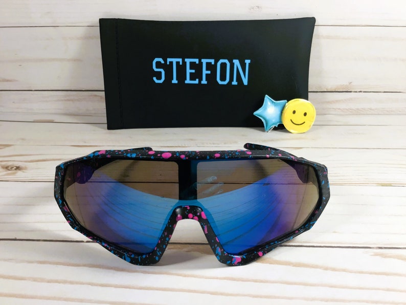 Puede incluir: Gafas de sol negras y rosas con manchas de pintura y lentes azules espejados. Las gafas de sol est&aacute;n sobre una superficie de madera con una funda negra que dice "STEFON" en letras azules.